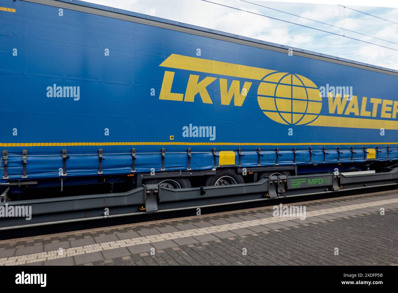Un container blu, marchiato con il logo „LKW Walter“, è parcheggiato su un carro su binari ferroviari. Ci sono linee elettriche aeree. Foto Stock
