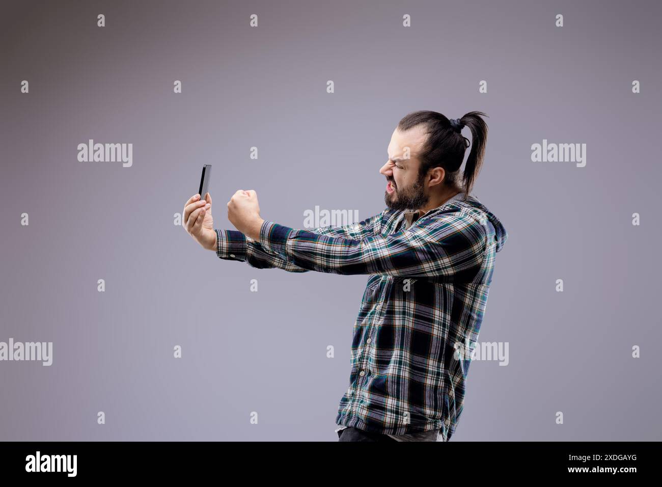 L'uomo barbuto con una coda di cavallo mostra emozioni intense mentre utilizza uno smartphone, riflettendo lo stress e il travolgimento dell'impatto della tecnologia sulla salute mentale Foto Stock