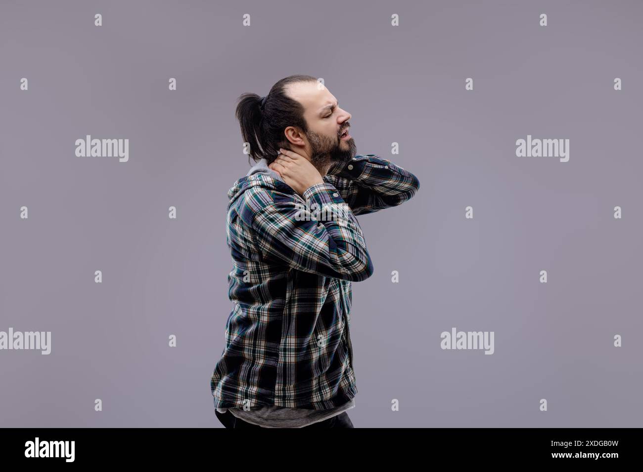 Uomo casual con barba e coda di cavallo che indossa una camicia a quadri, in piedi e soffre di dolore al collo, massaggiando il collo con un'espressione dolorosa Foto Stock