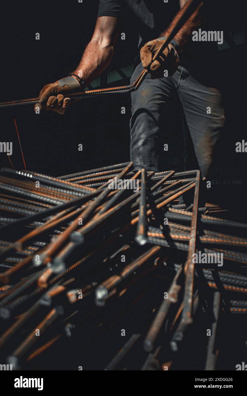 Un uomo non identificabile con camicia scura, jeans sporchi e guanti da lavoro si aggiunge a una pila di tondini in acciaio piegati all'interno di un magazzino buio. Foto Stock