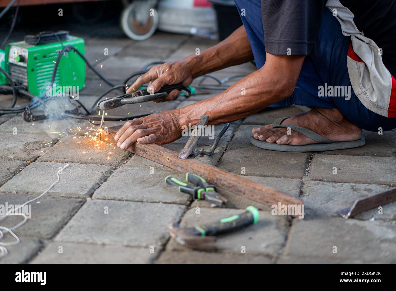 Una persona che salda il metallo con una saldatrice su una superficie pavimentata. Le scintille sono visibili e vari strumenti sono sparsi intorno. Foto Stock