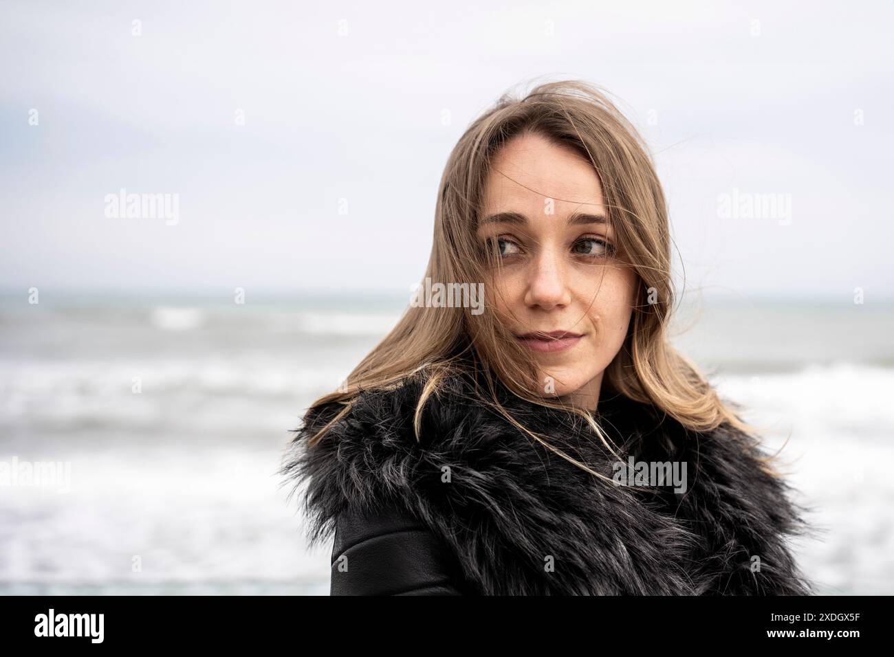 Donna con un cappotto di pelliccia che guarda con cura la spiaggia in una ventosa giornata invernale. Guarda in lontananza con il mare e le onde sullo sfondo, si conva Foto Stock