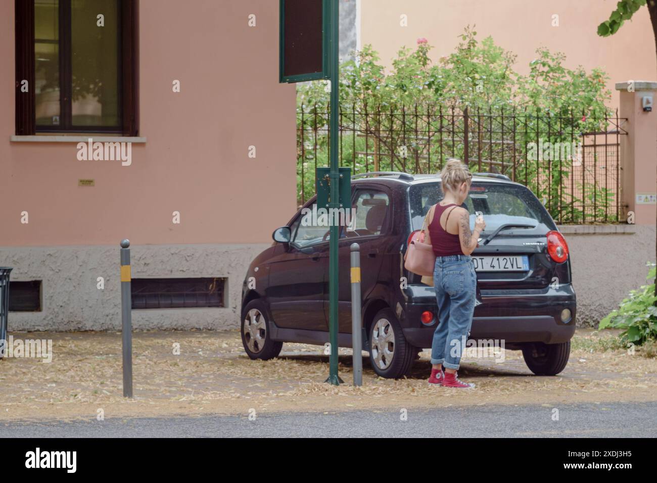 Cremona, Italia - 21 giugno 2024 donna in attesa alla fermata dell'autobus in una zona residenziale con alberi e un edificio sullo sfondo in una soleggiata estate Foto Stock
