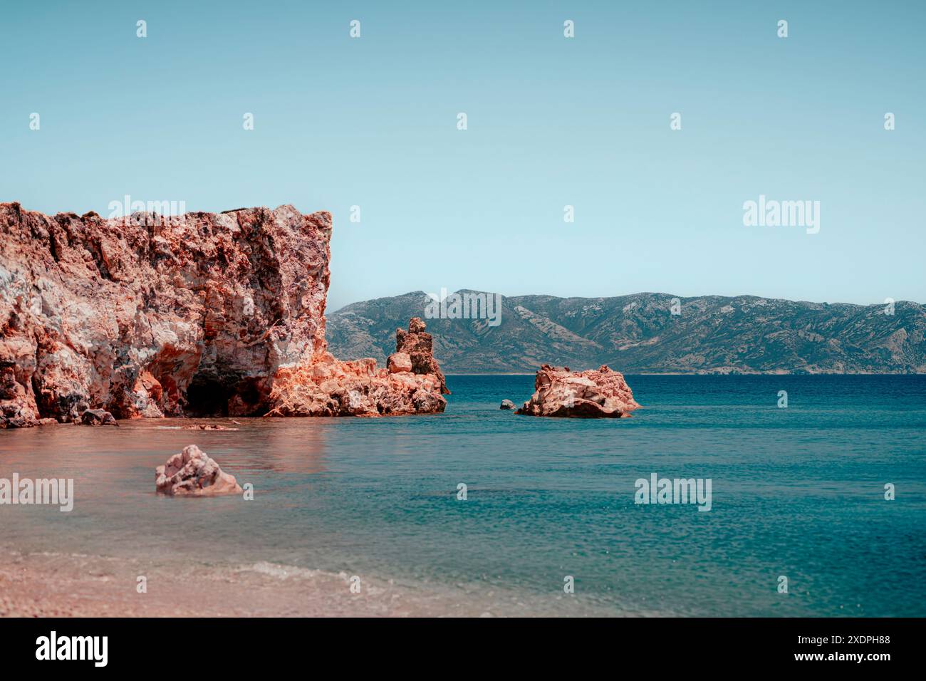 Vista di una spiaggia di sabbia rosa nell'isola di Milos, in Grecia, con acque cristalline e montagne sullo sfondo durante una soleggiata giornata estiva Foto Stock