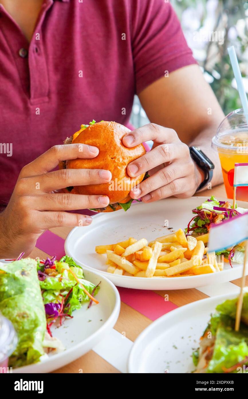 Man Boy che tiene in mano un hamburger di pollo fritto glassato al miele in panino brioche servito con patatine fritte e insalata su un piatto in un ambiente caffetteria. Foto Stock