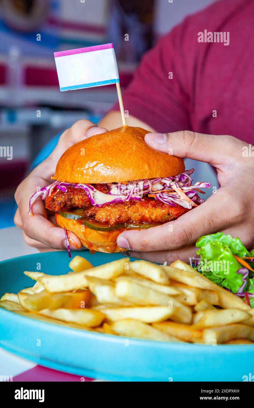 Man Boy che tiene in mano un hamburger di pollo fritto glassato al miele in panino brioche servito con patatine fritte e insalata su un piatto in un ambiente caffetteria. Foto Stock