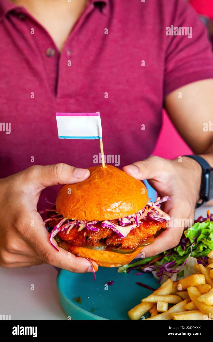 Man Boy che tiene in mano un hamburger di pollo fritto glassato al miele in panino brioche servito con patatine fritte e insalata su un piatto in un ambiente caffetteria. Foto Stock