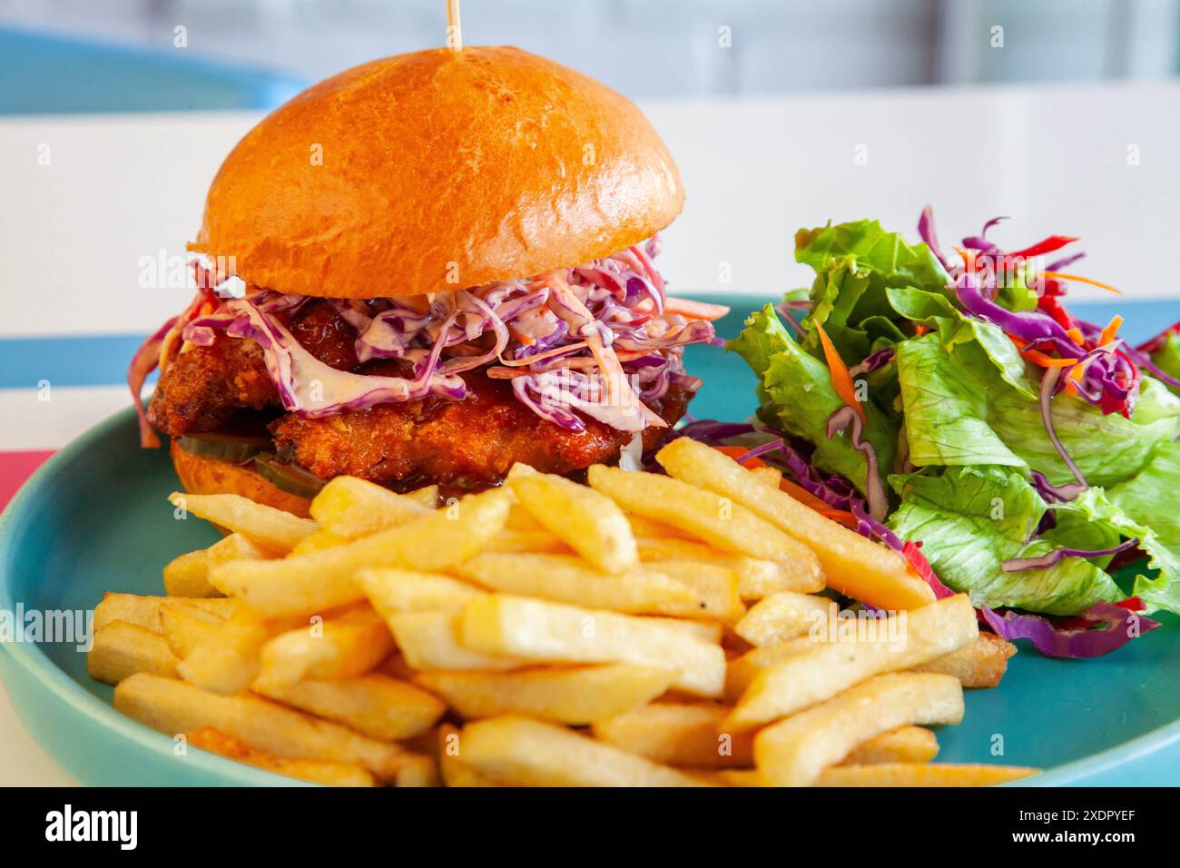 Hamburger di pollo fritto glassato al miele in panino brioche servito con patatine fritte e insalata su un piatto in un ambiente caffetteria. Foto Stock