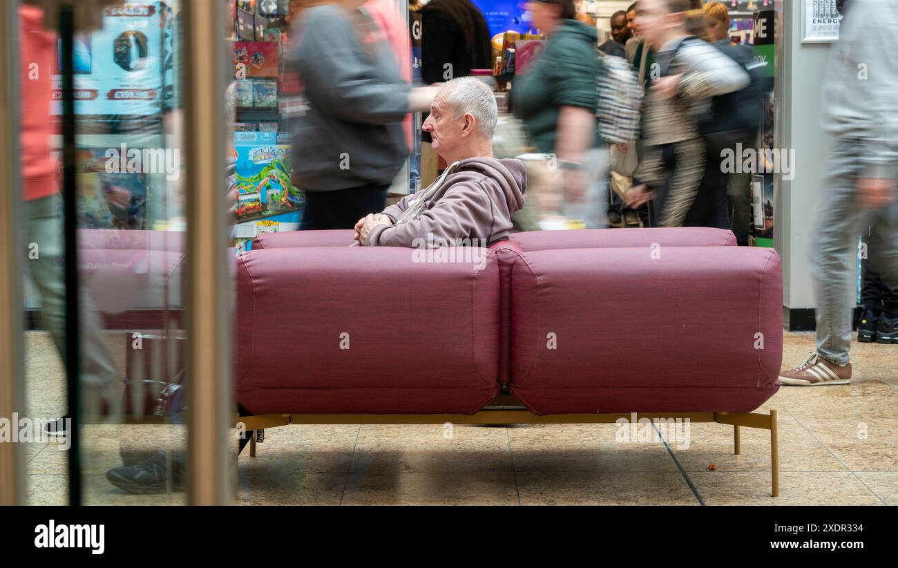 Foto a velocità dell'otturatore lunga di un uomo anziano seduto a spianare su un posto in un centro commerciale, con gli acquirenti che si muovono intorno e dietro di lui Foto Stock
