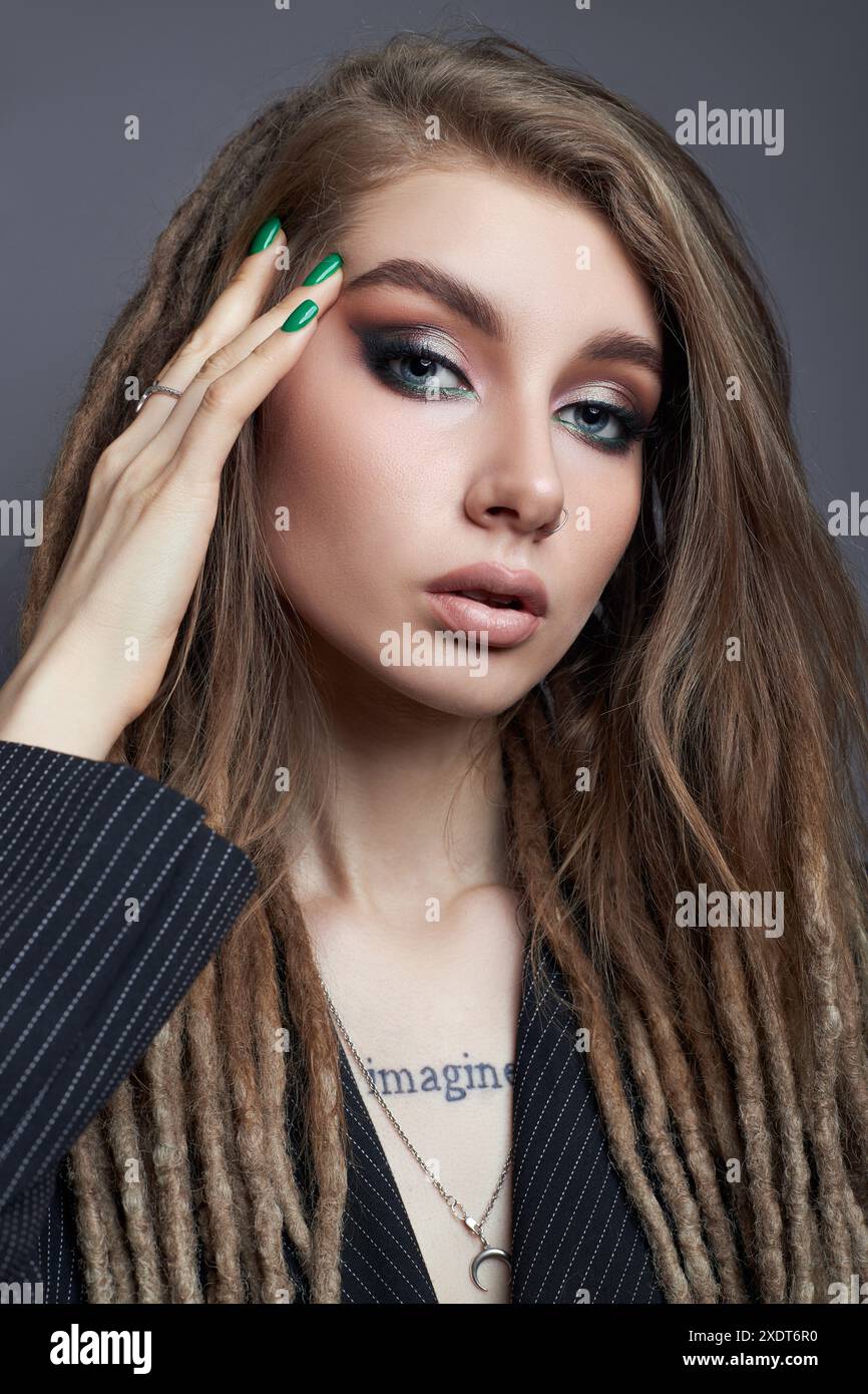 Beaity Woman con smalto verde e lunghi dreadlock marroni posa per un ritratto in un ambiente da studio, indossando un blazer nero con un pinstripe bianco Foto Stock
