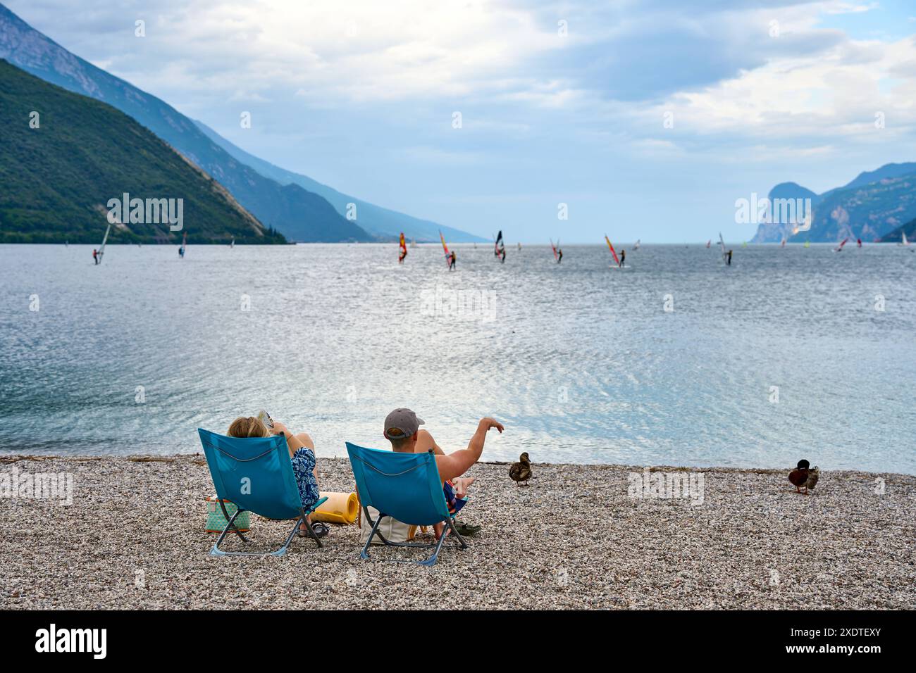 Torbole, Lago di Garda, Italia - 24 giugno 2024: due turisti in coppia su una sdraio sulla spiaggia del Lago di Garda in Italia guardando il panorama con windsurf e montagne *** Zwei Touristen als Pärchen in einem Liegestuhl am Strand vom Gardasee in Italien blicken auf das Panorma mit Windsurfern und Bergen Foto Stock