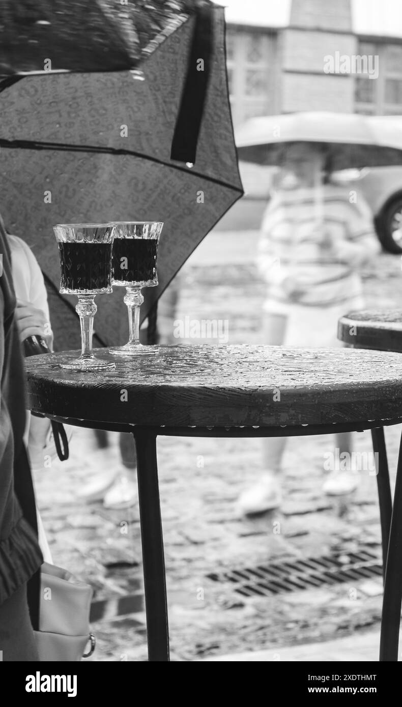 due bicchieri di vino rosso sul tavolo sotto la pioggia, monocromatico. Appuntamento sotto la pioggia. Bicchieri da vino sotto l'ombrello nel caffè sul marciapiede. Pioggia intensa in città. Foto Stock