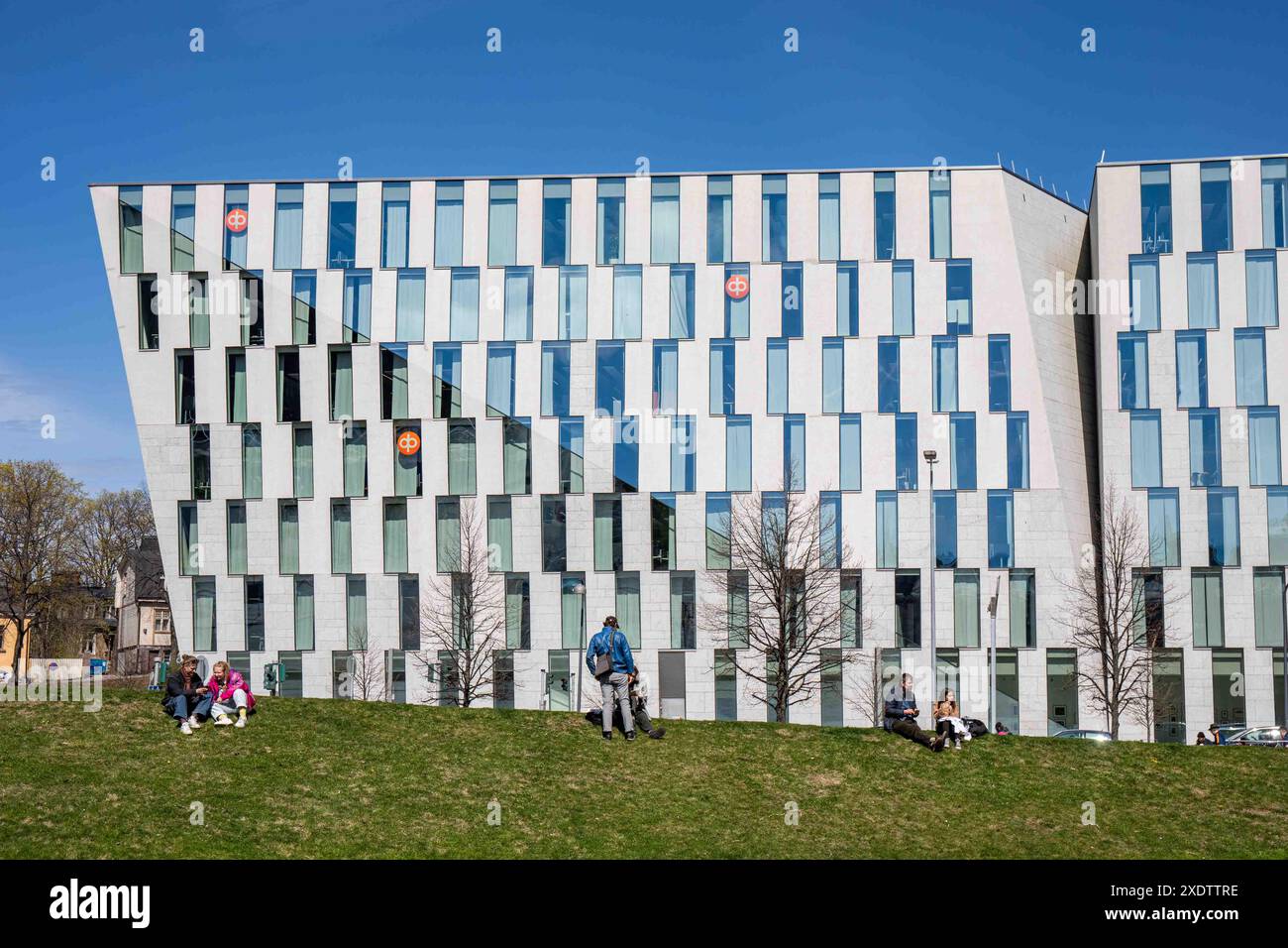 Persone che si godono una giornata di primavera soleggiata sul prato knoll nel Parco Dallapé sullo sfondo della sede centrale del gruppo OP nel quartiere Vallila di Helsinki, Finlandia. Foto Stock