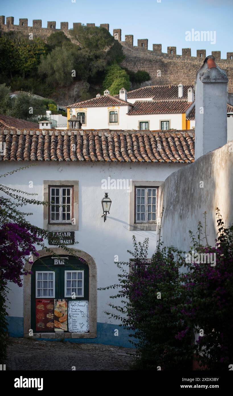 Rua de São Teotónio, Óbidos, Portogallo. Stretta strada acciottolata che conduce ad un ristorante in un tradizionale edificio medievale a due piani con tetto in cotto, dipinto di bianco. La sezione settentrionale del muro di calcare che circonda il villaggio sullo sfondo. Foto Stock
