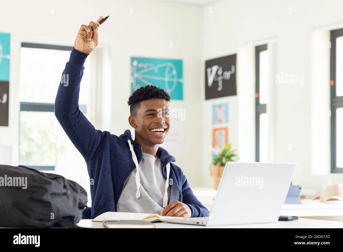 A scuola, uno studente maschio che alza la mano mentre usa il laptop in classe, sorridendo Foto Stock