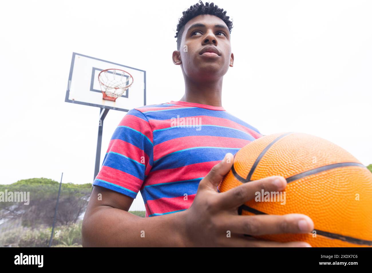 Tiene in mano pallacanestro, ragazzo afroamericano adolescente sul campo all'aperto, sembra determinato Foto Stock