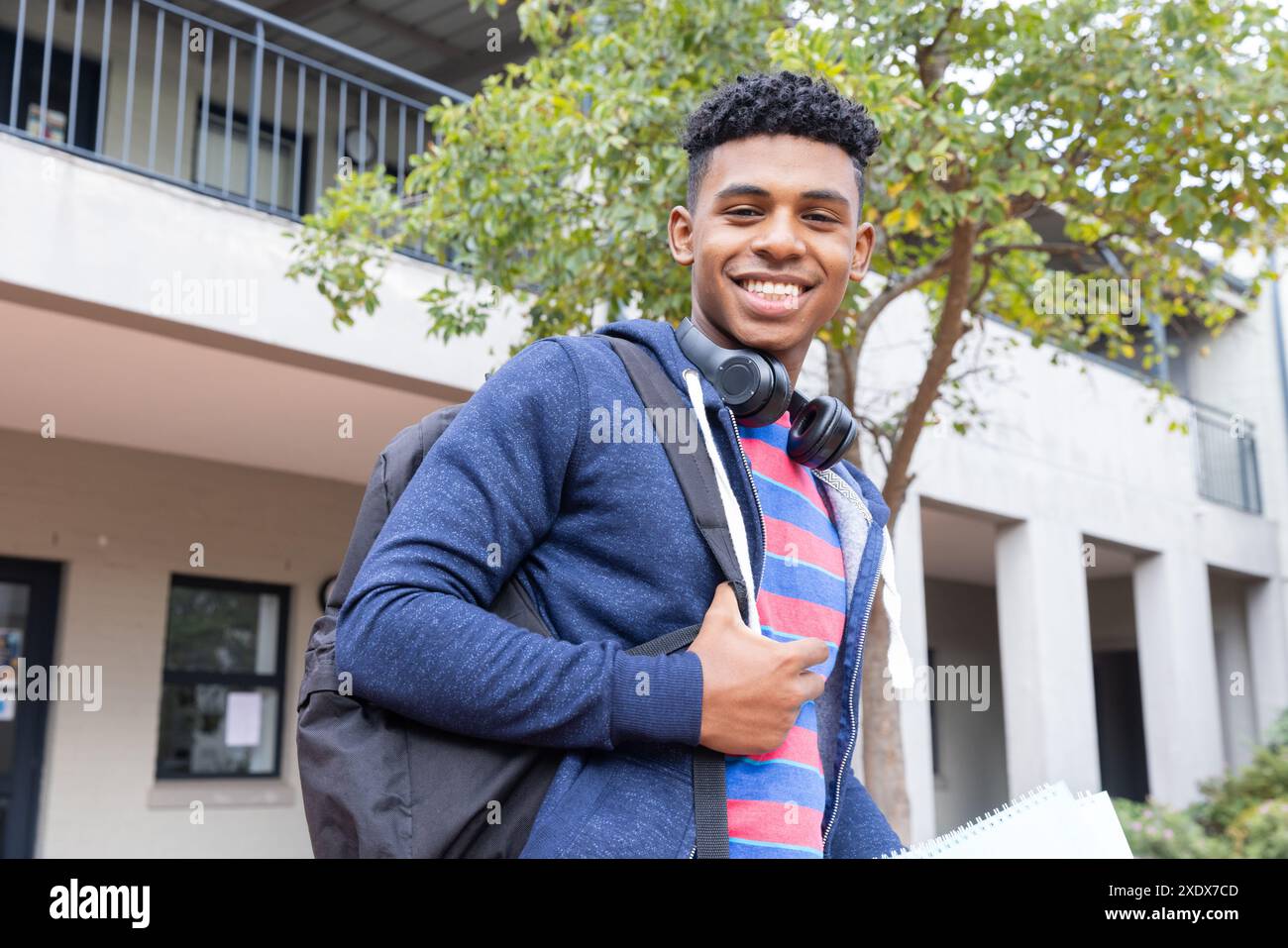 Studente maschio sorridente con zaino e cuffie in piedi fuori dall'edificio scolastico Foto Stock