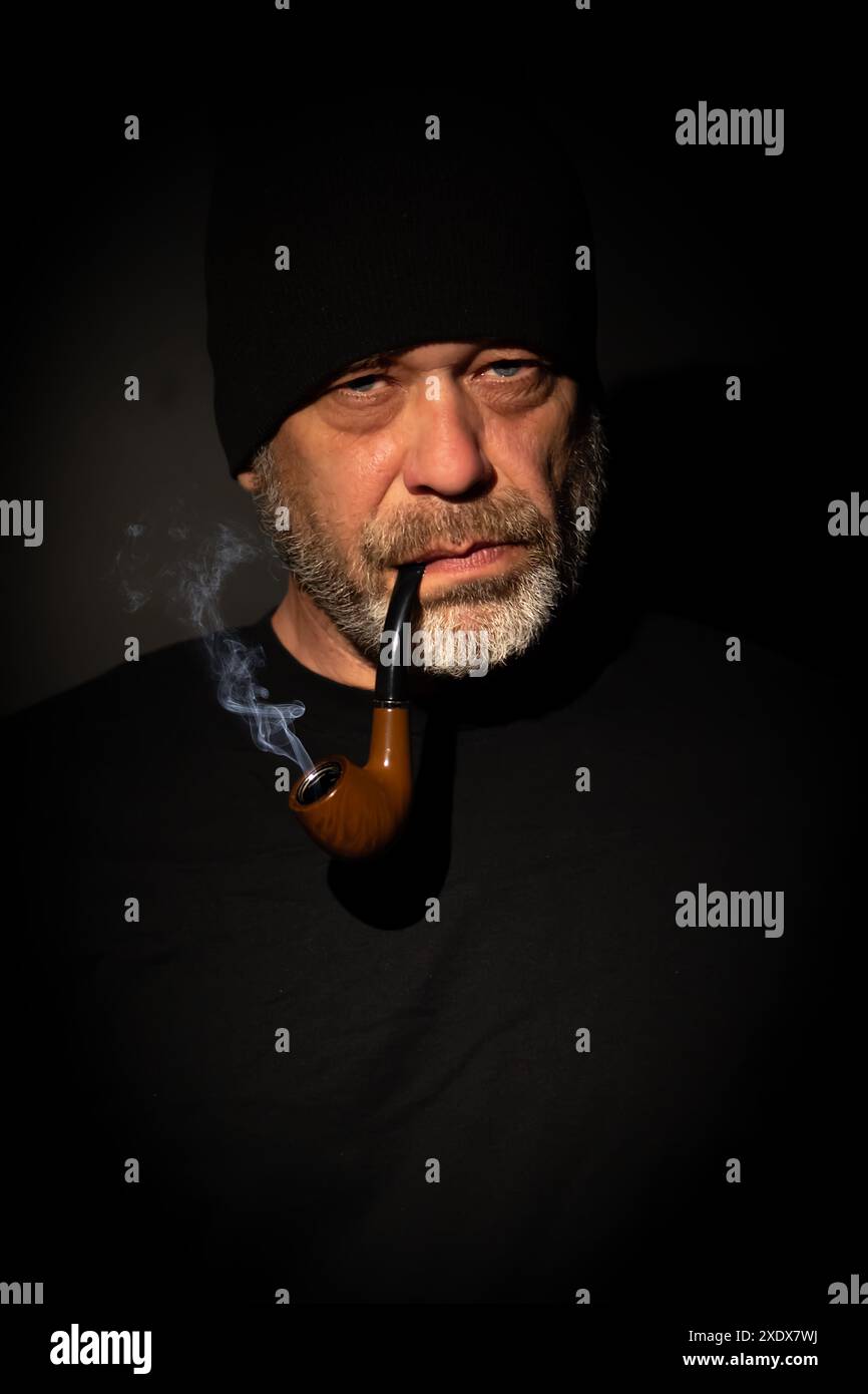 Un uomo anziano con la barba grigia che indossa un cappello nero e una camicia nera fuma una pipa, tonalità scura. Il fumo si arriccia verso l'alto, l'espressione del viso è se Foto Stock