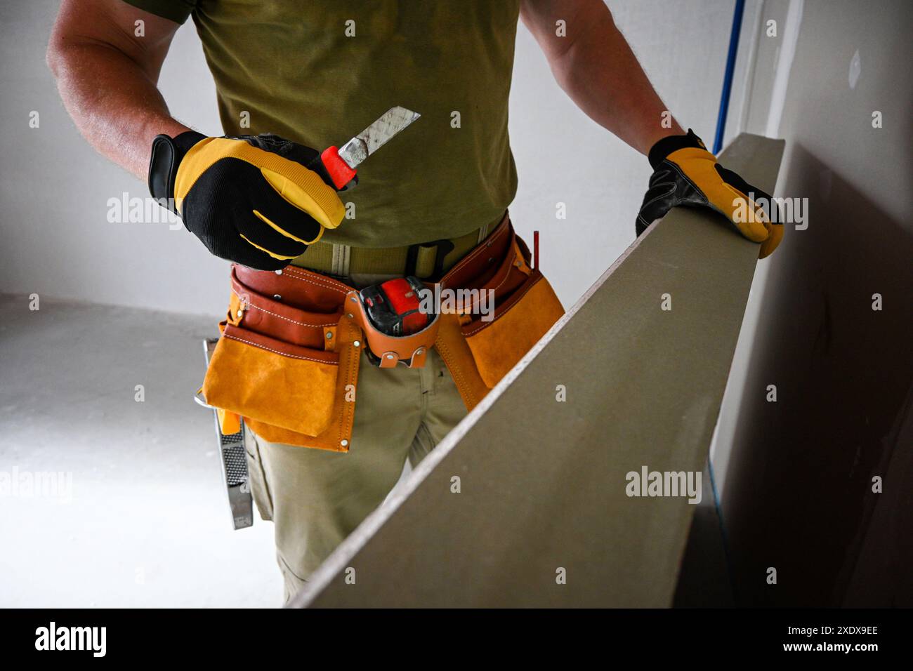 Il lavoratore edile indossa la cintura portautensili e tiene in mano la cartongesso e il coltello per usi generici che si prepara al taglio. Foto Stock