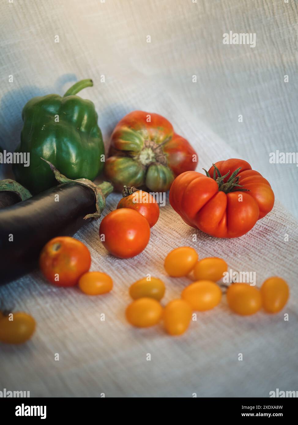 Natura morta verticale di pomodori, melanzane e peperone su un panno di cotone bianco alla luce del sole dorato Foto Stock