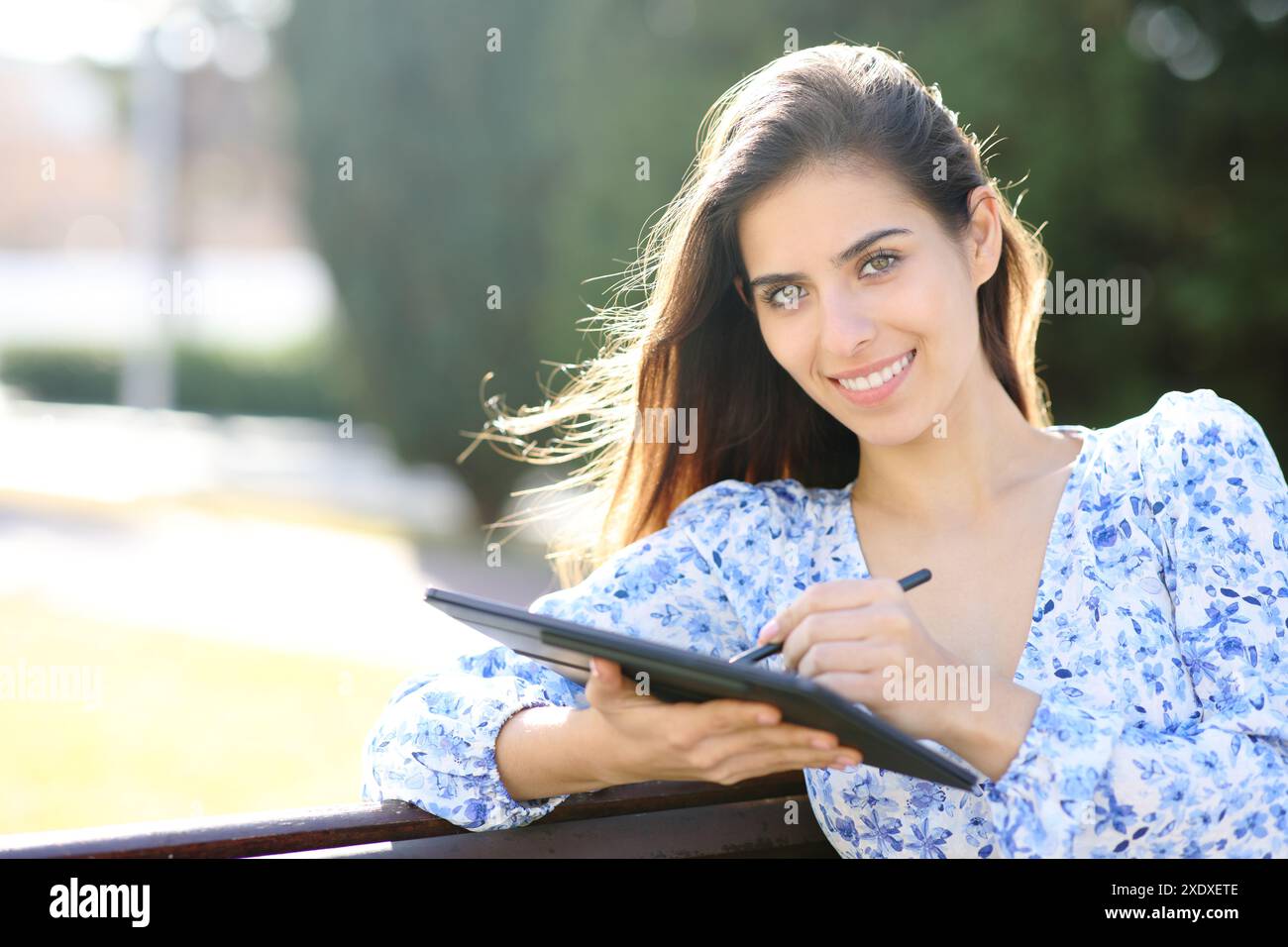 Una donna felice che disegna su un tablet con la penna ti guarda in un parco Foto Stock