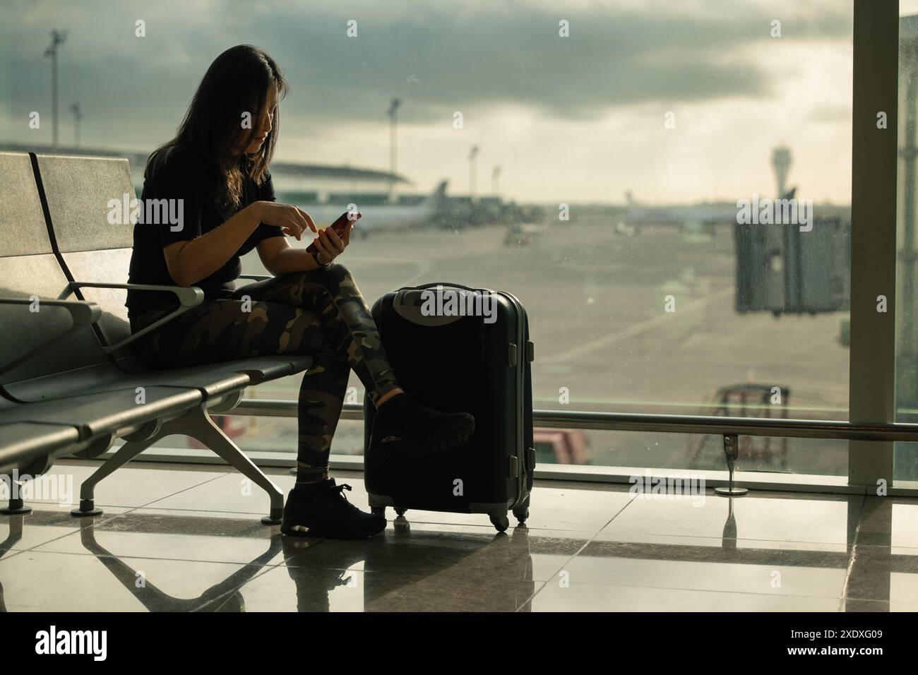 Una donna controlla il suo telefono cellulare nell'area di imbarco dell'aeroporto e accanto a lei una finestra con l'area esterna dell'aeroporto di ​​the. Foto Stock