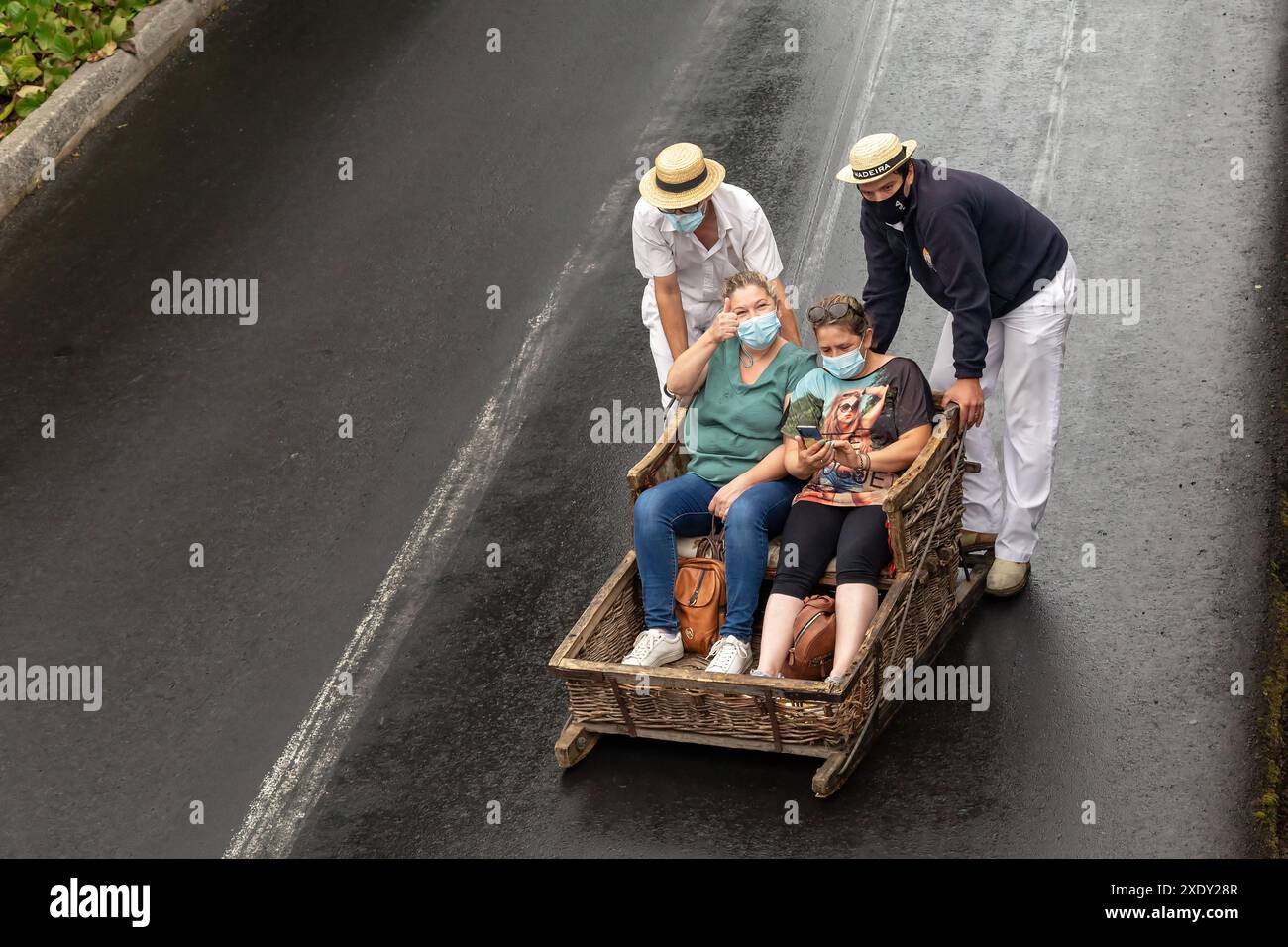 FUNCHAL, PORTOGALLO - 24 AGOSTO 2021: Donne non identificate corrono felicemente per strada in cesti di vimini (slitte) con l'aiuto di autisti tradizionali Foto Stock