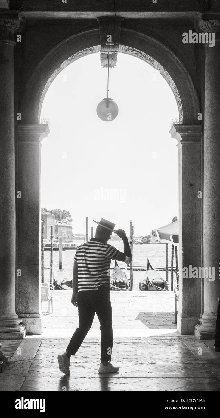 Venezia, Italia - 30 giugno 20220 gondoliere che si allontana dalla telecamera sotto un arco con gondole sullo sfondo Foto Stock
