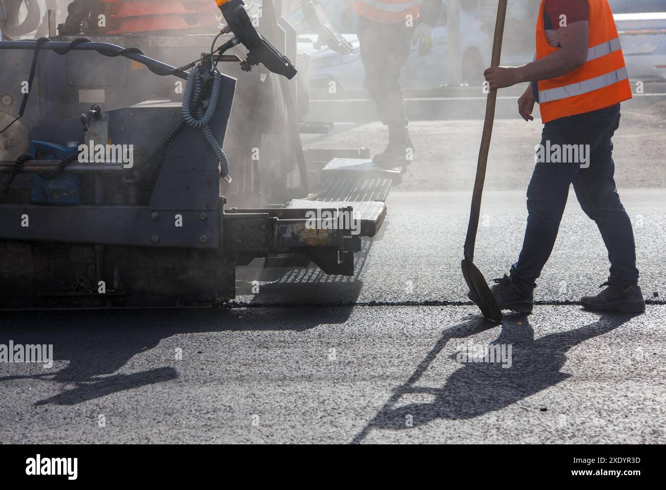Gli uomini che lavorano con l'asfaltatrice durante la riparazione stradale lavorano alla luce del giorno con fumo e vapore nell'aria Foto Stock