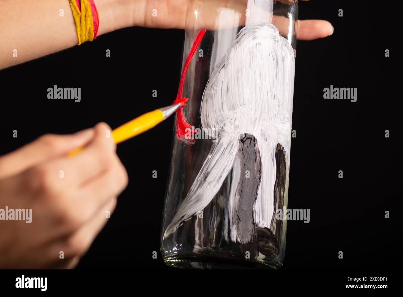 Un artista che tiene e dipinge una bottiglia di vetro con vernice gouache rossa e nera. Isolato su sfondo nero. Foto Stock