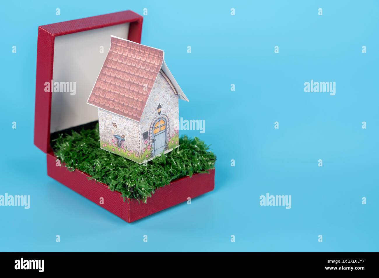 Una piccola casa è collocata all'interno di una scatola rossa. La casa è fatta di carta e si trova su un'area verde erbosa. La scatola è rossa e ha una finitura lucida. Il Foto Stock