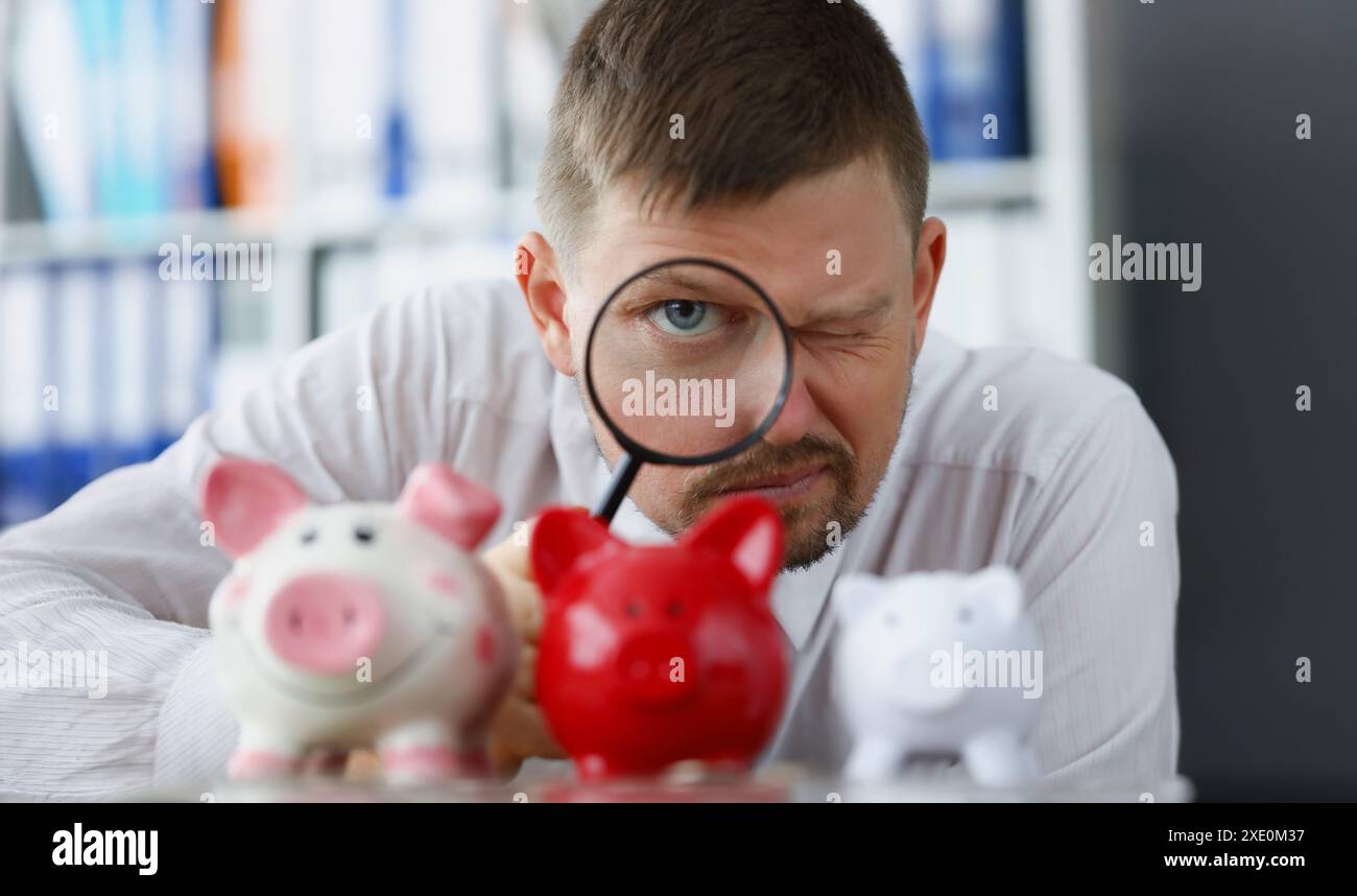 L'uomo guarda attraverso la lente d'ingrandimento al salvadanaio. Ricerca di depositi e investimenti redditizi Foto Stock