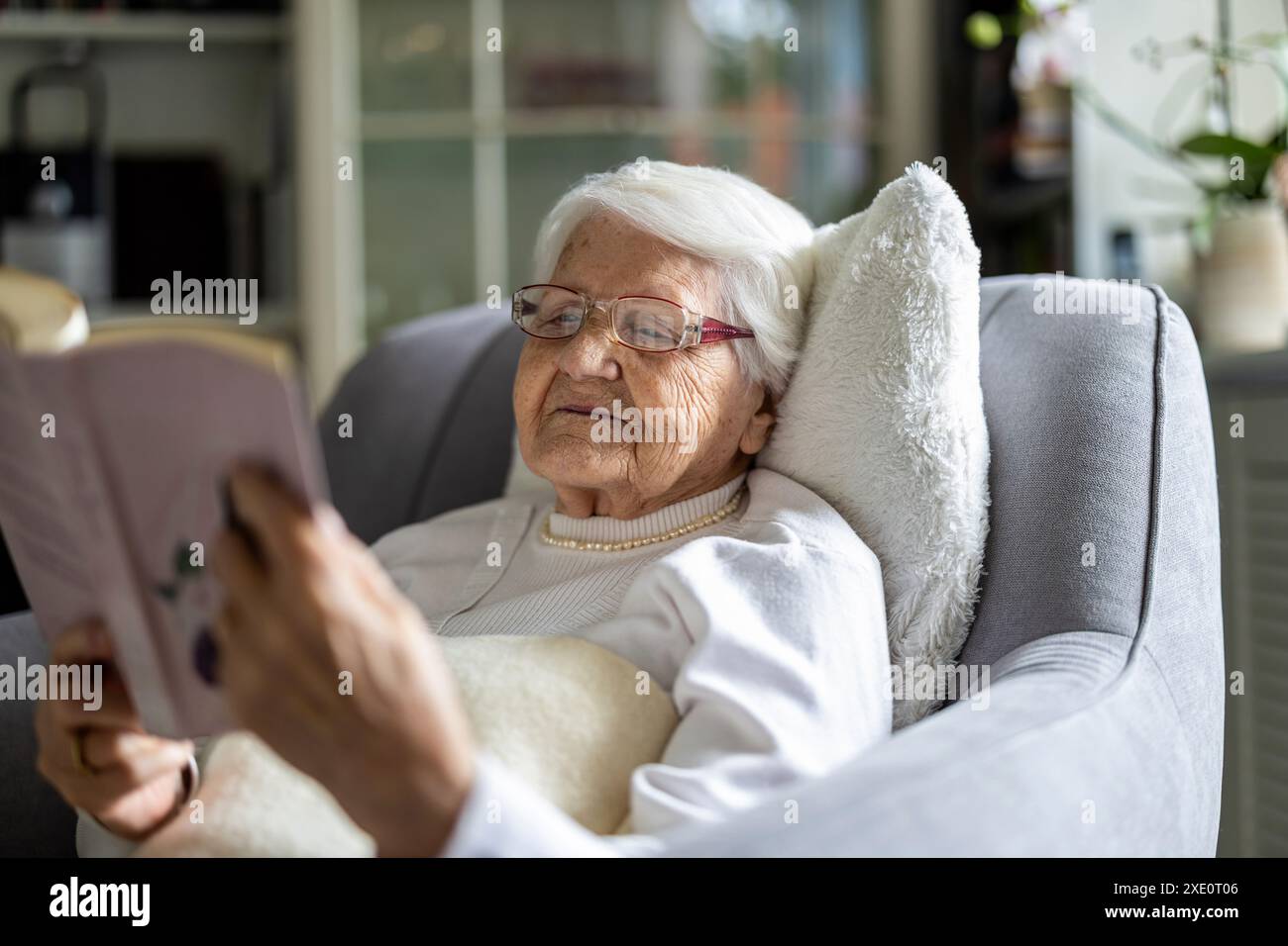 Donna anziana che legge un libro in una comoda poltrona a casa Foto Stock