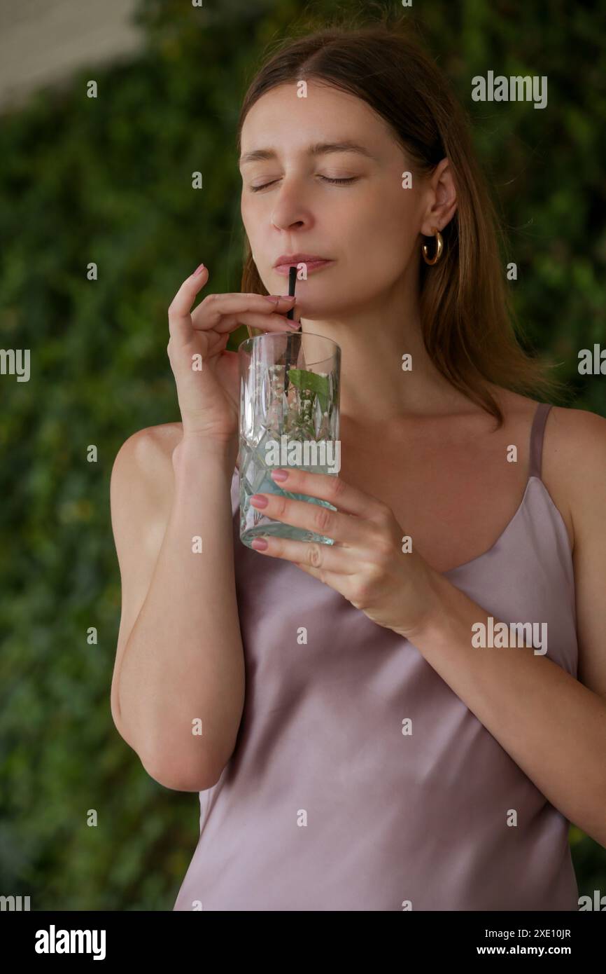 Elegante donna in abito camisole di seta con un bicchiere di cocktail mojito appena preparato e godendosi una tranquilla giornata estiva Foto Stock
