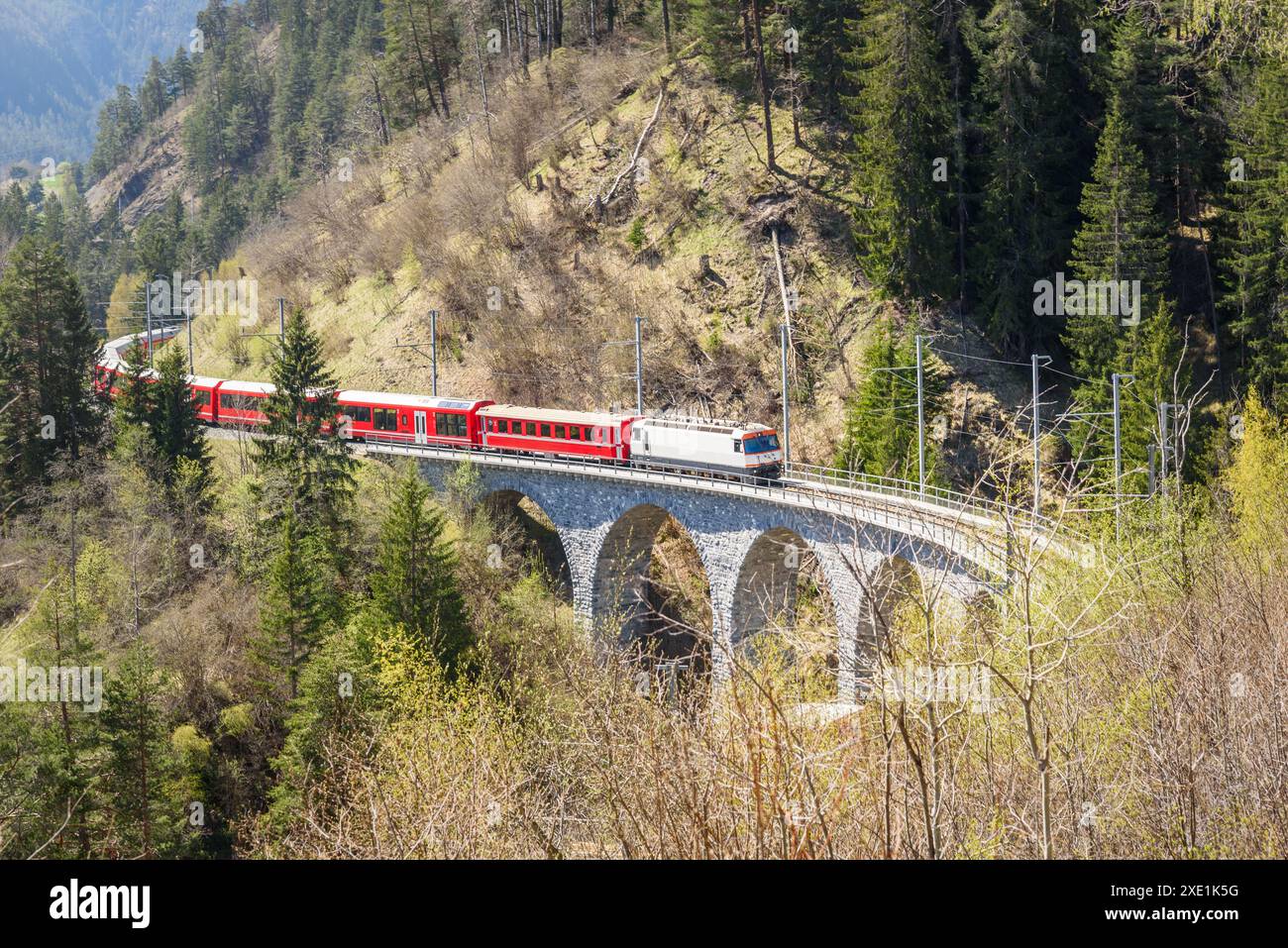 Treno elettrico passeggeri che attraversa un viadotto in pietra ondulato sulle montagne in una giornata di primavera assolata Foto Stock