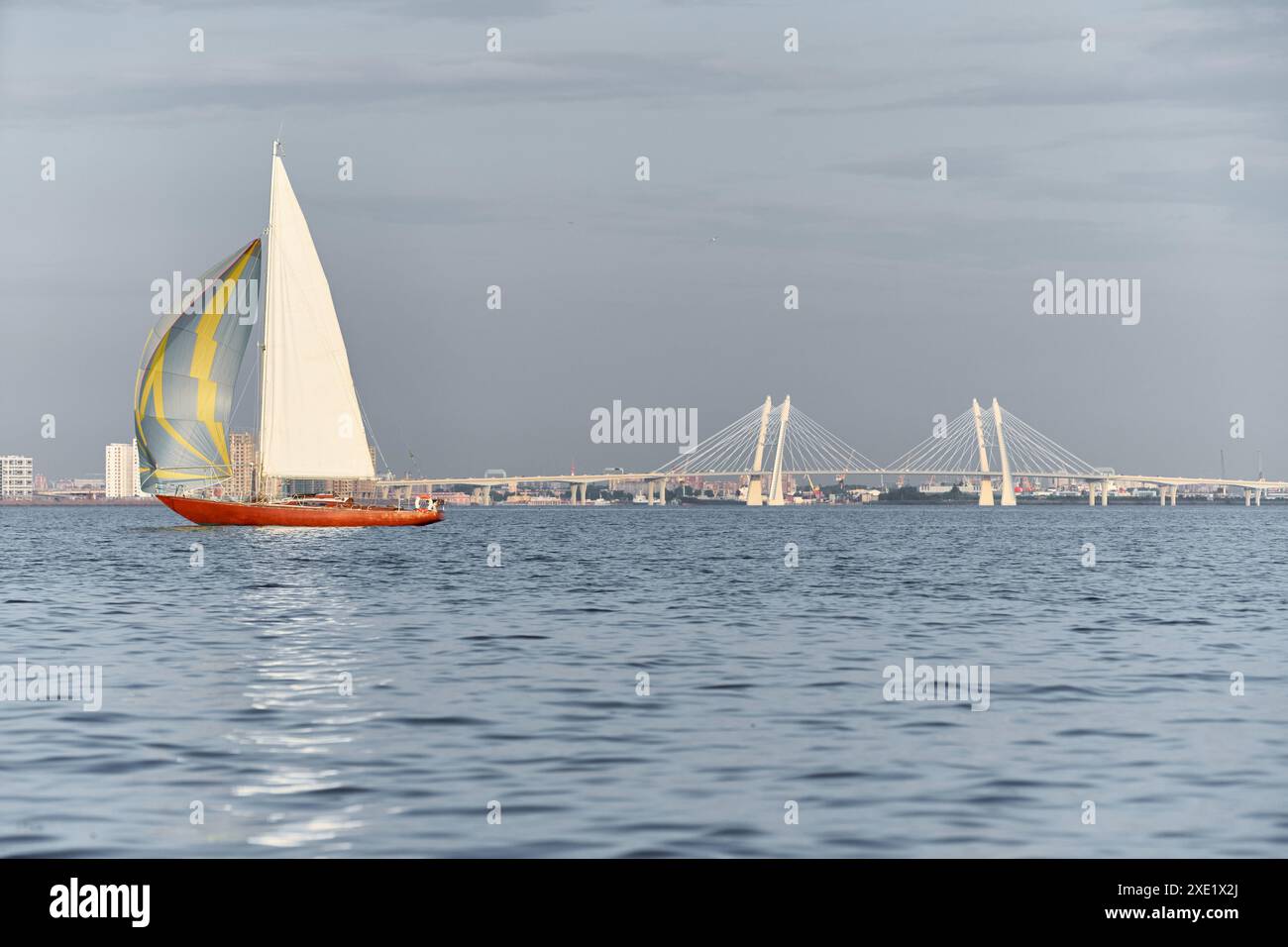 Classico yacht a vela in mare al tramonto, ponte strallato sullo sfondo, cielo limpido, riflesso del sole sull'acqua Foto Stock Classico yacht a vela in mare al tramonto, ponte strallato sullo sfondo, cielo limpido, riflesso del sole sull'acqua Foto Stock