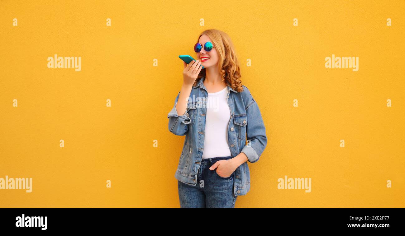 Giovane donna felice che tiene la conversazione al telefono, utilizza un registratore a comando vocale, un assistente o risponde alle chiamate sullo smartphone indossando abiti casual su un luminoso YE Foto Stock