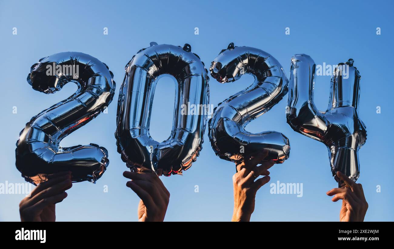 Palloncino Silver foil numero 2024 per festeggiare l'anno nuovo su sfondo blu. Concetto di auguri di buon anno. Mani che tengono Bal Foto Stock