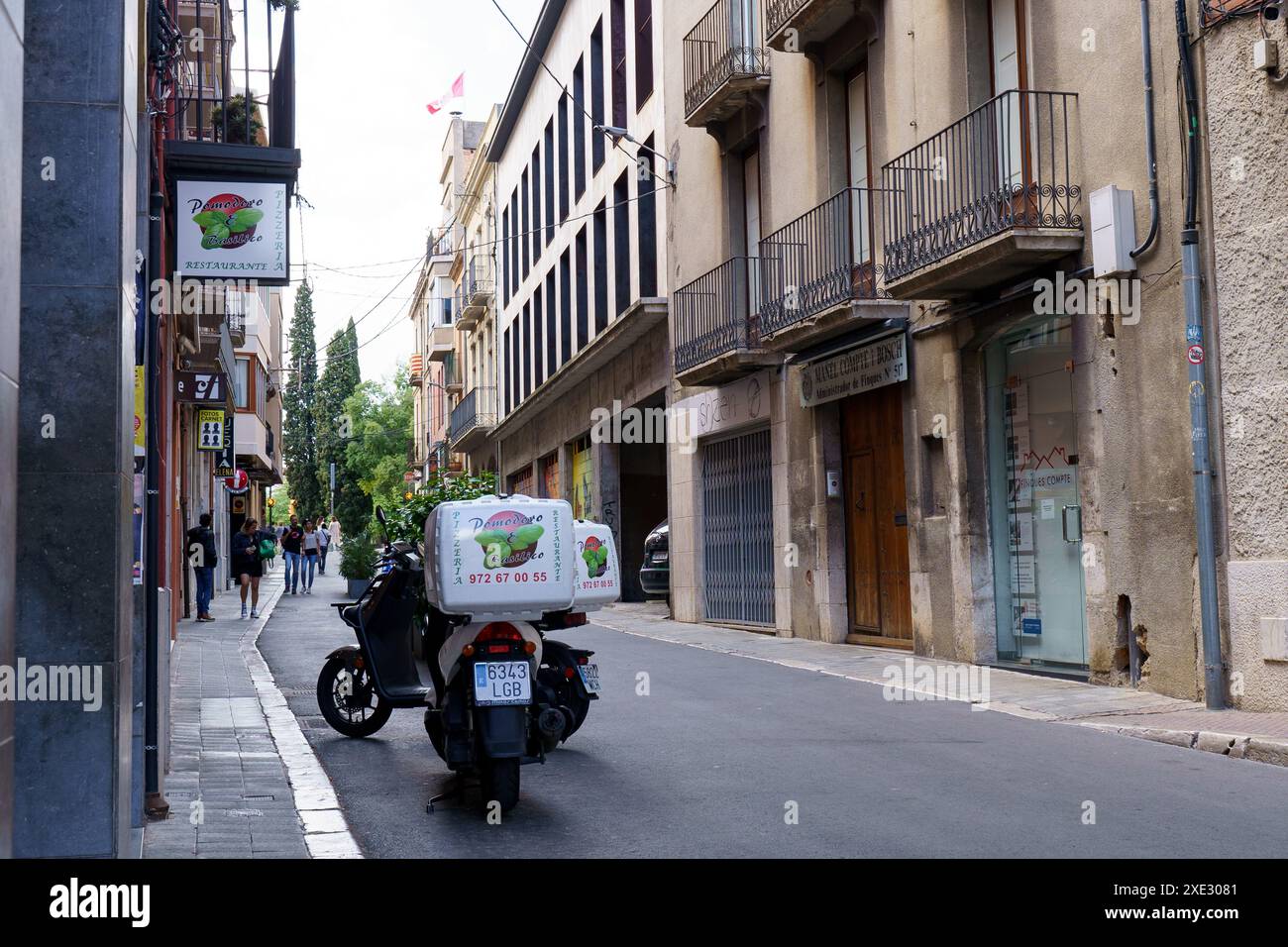 Figueres, Spagna - 14 maggio 2023: Uno scooter per consegne accelera lungo una strada pittoresca e stretta fiancheggiata da edifici tradizionali e cartelli Foto Stock