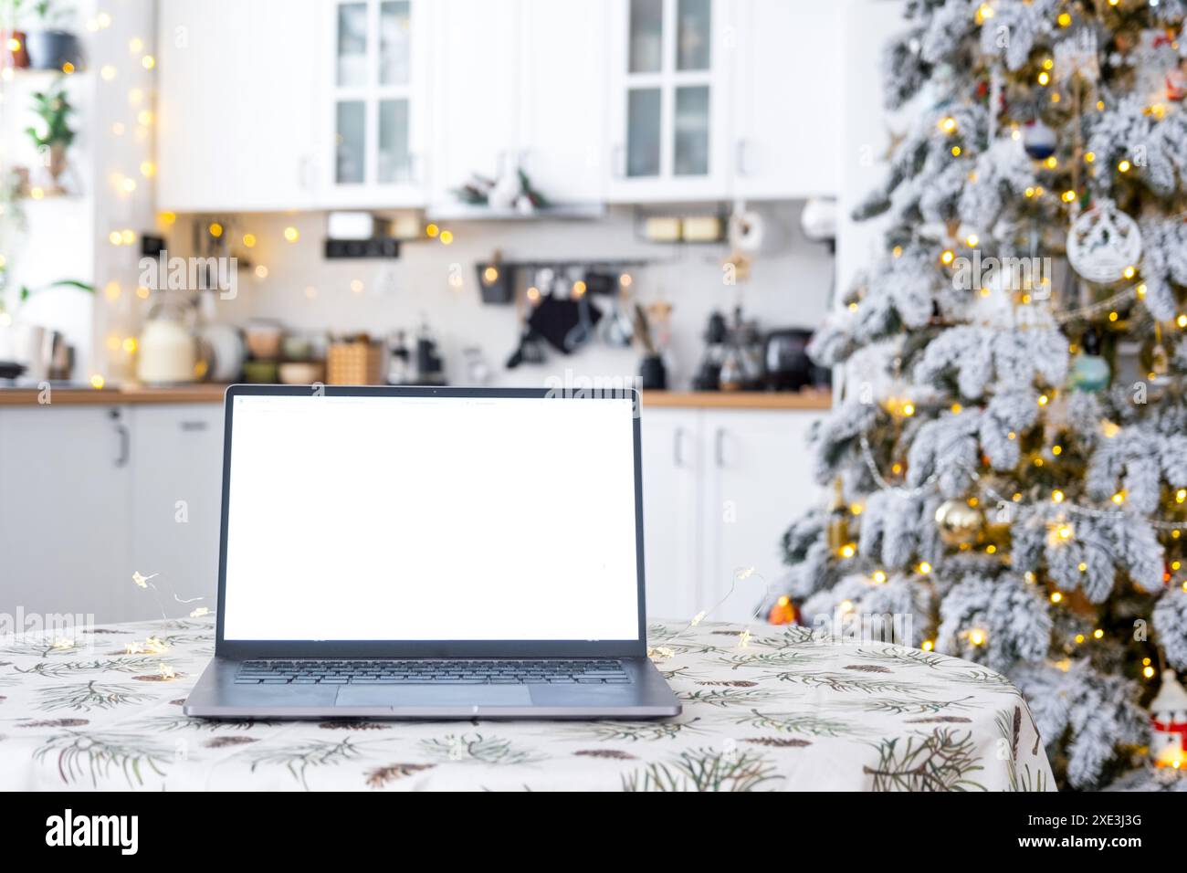 Computer portatile con schermo bianco ripreso nell'accogliente cucina di Natale decorata di colore bianco con luci delle fate e un albero di Natale. Seasona Foto Stock