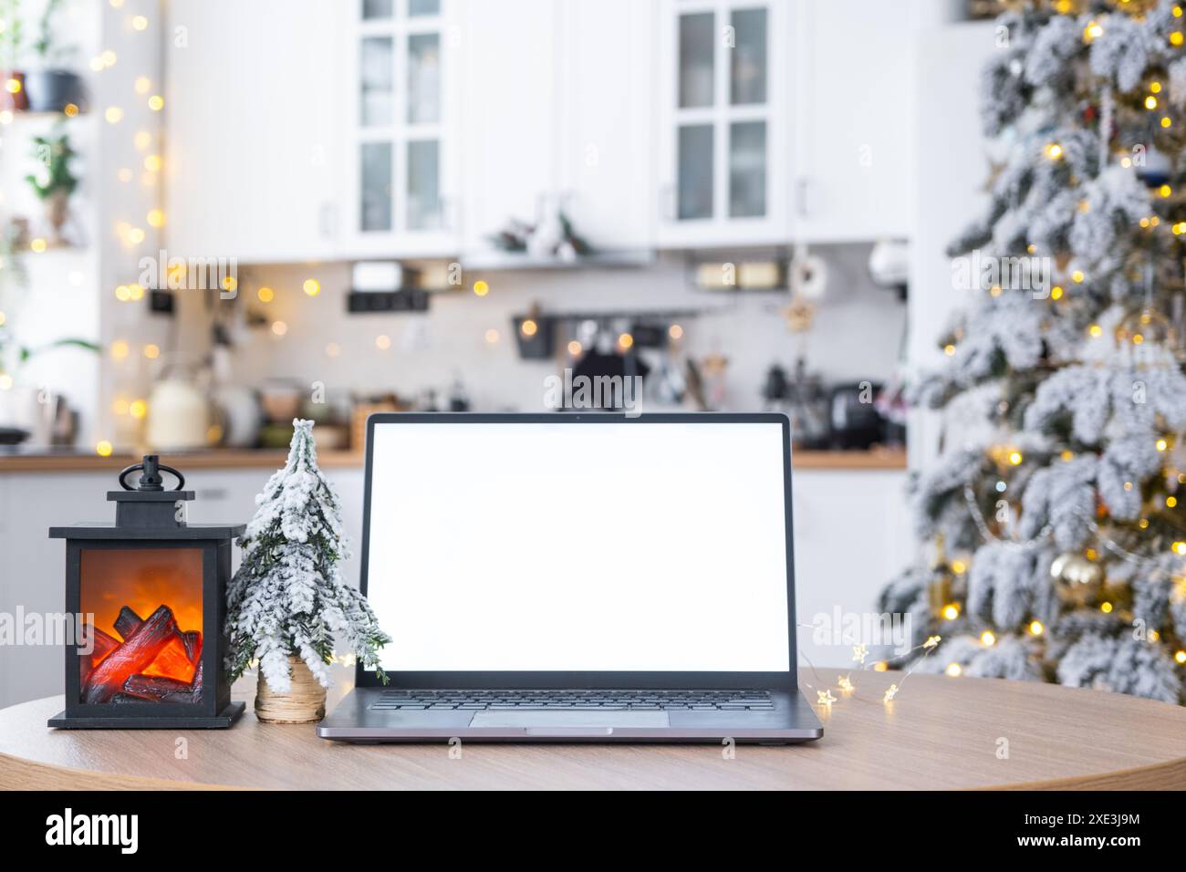 Computer portatile con schermo bianco ripreso nell'accogliente cucina di Natale decorata di colore bianco con luci delle fate e un albero di Natale. Seasona Foto Stock