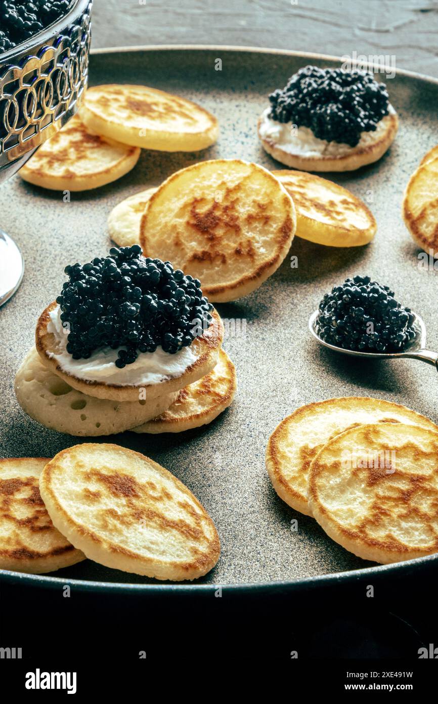 Blinis con caviale nero, su un piatto festivo su sfondo nero, mini pancake Foto Stock