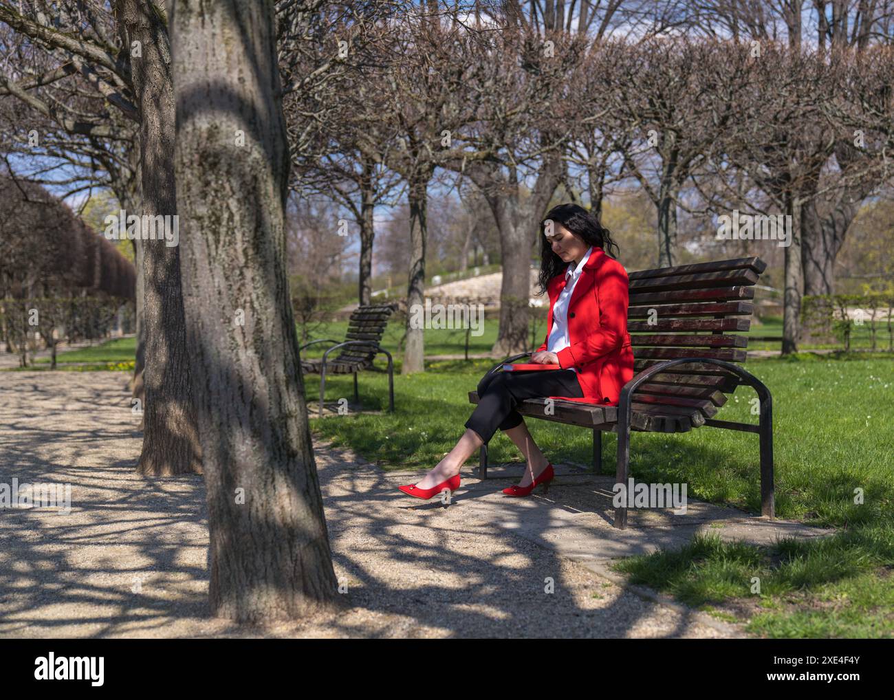 Un'elegante donna d'affari siede su una panchina del parco e scrive su un notebook. La signora indossa un mantello rosso, una camicetta bianca e scarpe rosse. Foto Stock