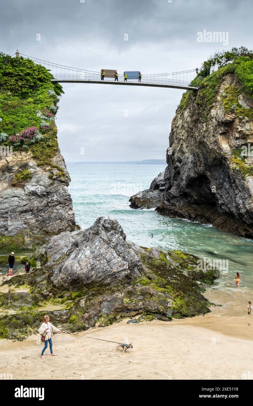 I vacanzieri si rilassano e si divertono in mare e sulla spiaggia in riva al mare mentre gli operai attraversano il ponte sospeso sopra la spiaggia di Towan a New Foto Stock