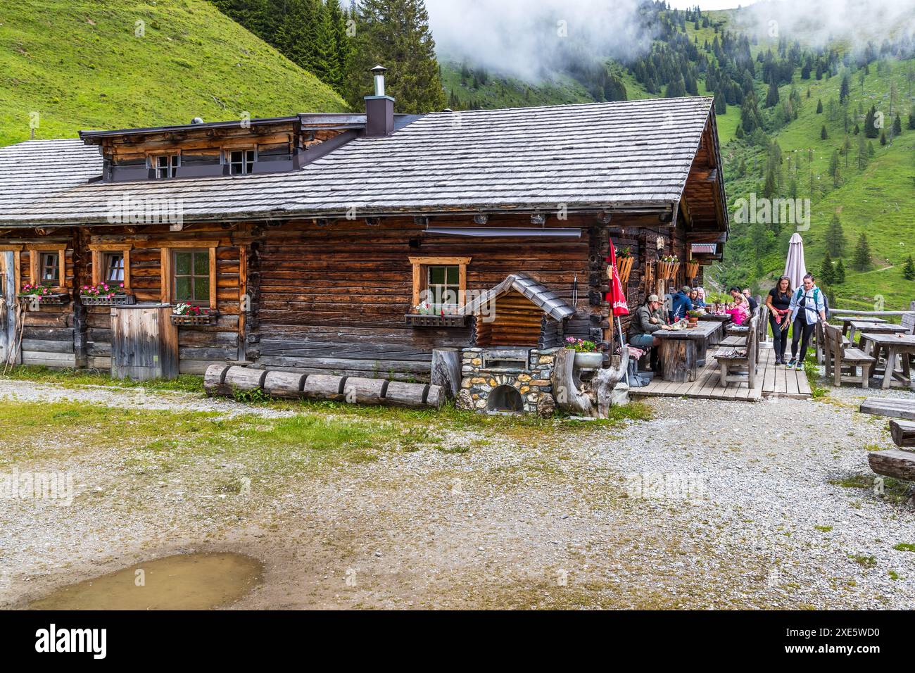 Gli ospiti sono seduti sulla terrazza di una baita alpina con un frontone in legno scuro lungo il sentiero Salzburger Almenweg vicino a Langbrand nella valle di Großarl, circondati da banchi di nebbia sulle montagne, Salisburgo, Austria. Una bandiera rossa e bianca e un ombrellone si trovano all'ingresso. Foto Stock