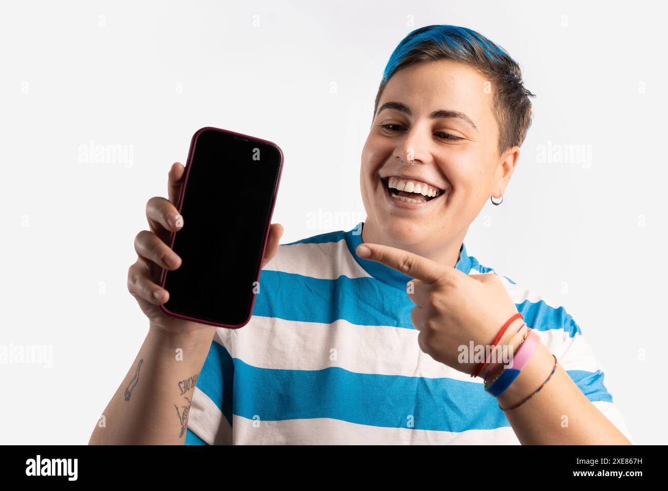 Una persona androgina con i capelli blu, indossa una camicia a righe blu e bianca e un bracciale Pride, sorride e punta allo schermo di uno smartphone, studio Foto Stock