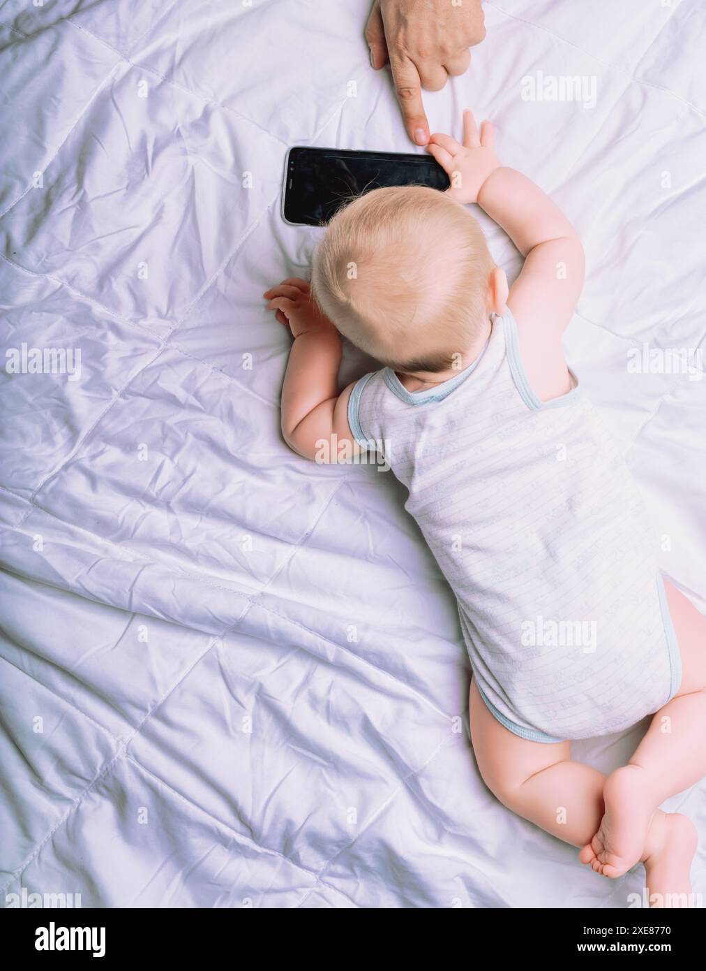 Il bambino giace su una trapunta bianca, raggiungendo uno smartphone guidato dalla mano di un adulto. Questa scena incarna il concetto di istruzione come guida attraverso la nuova c Foto Stock