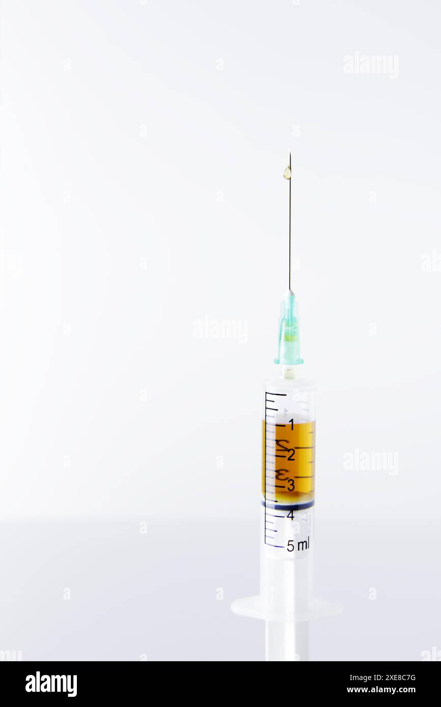 Primo piano della parte superiore di una siringa, con una goccia sull'ago, in posizione verticale su un tavolo da laboratorio bianco, con la dose di farmaco all'interno. Sfondo bianco Foto Stock
