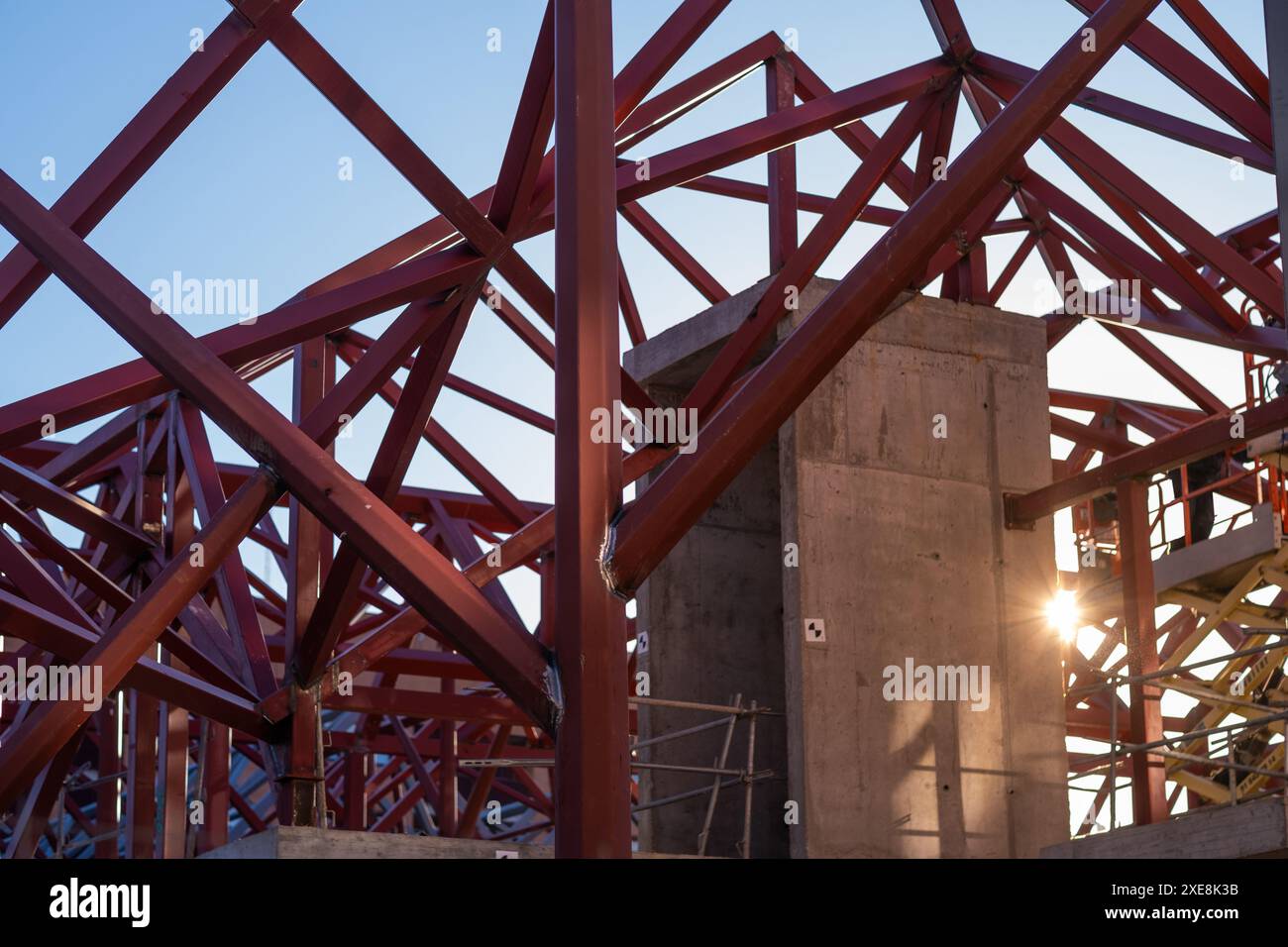 Composizione geometrica di travi metalliche rosse e gru blu in una costruzione moderna contro un cielo limpido. È l'alba e il sole entra tra la S Foto Stock