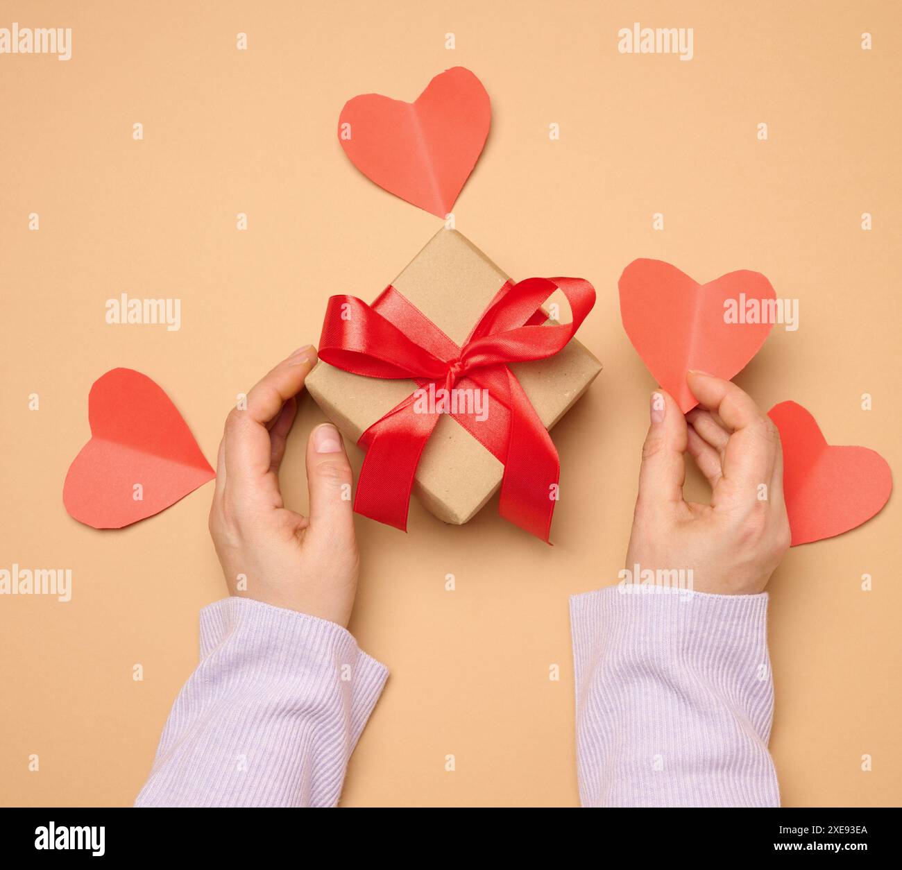 Una donna tiene in mano una confezione regalo avvolta da un nastro di seta rosso su sfondo beige Foto Stock